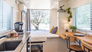 A compact kitchen with a coffee maker, a small dining area with a table and chairs, and a view of a tree outside. - Honeycomb Container Home | "Pickleball Court!" (Waco)