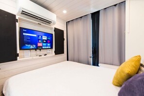 A hotel room with a bed, a mounted TV displaying various channels, an air conditioning unit, and a sliding door with curtains. - Honeycomb Container Home | "Pickleball Court!" (Waco)