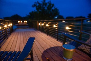 A wooden deck with a table and chairs, string lights, and a glass of beer. - "The Woods" Container | Pickleball | Rooftop Patio (Waco)