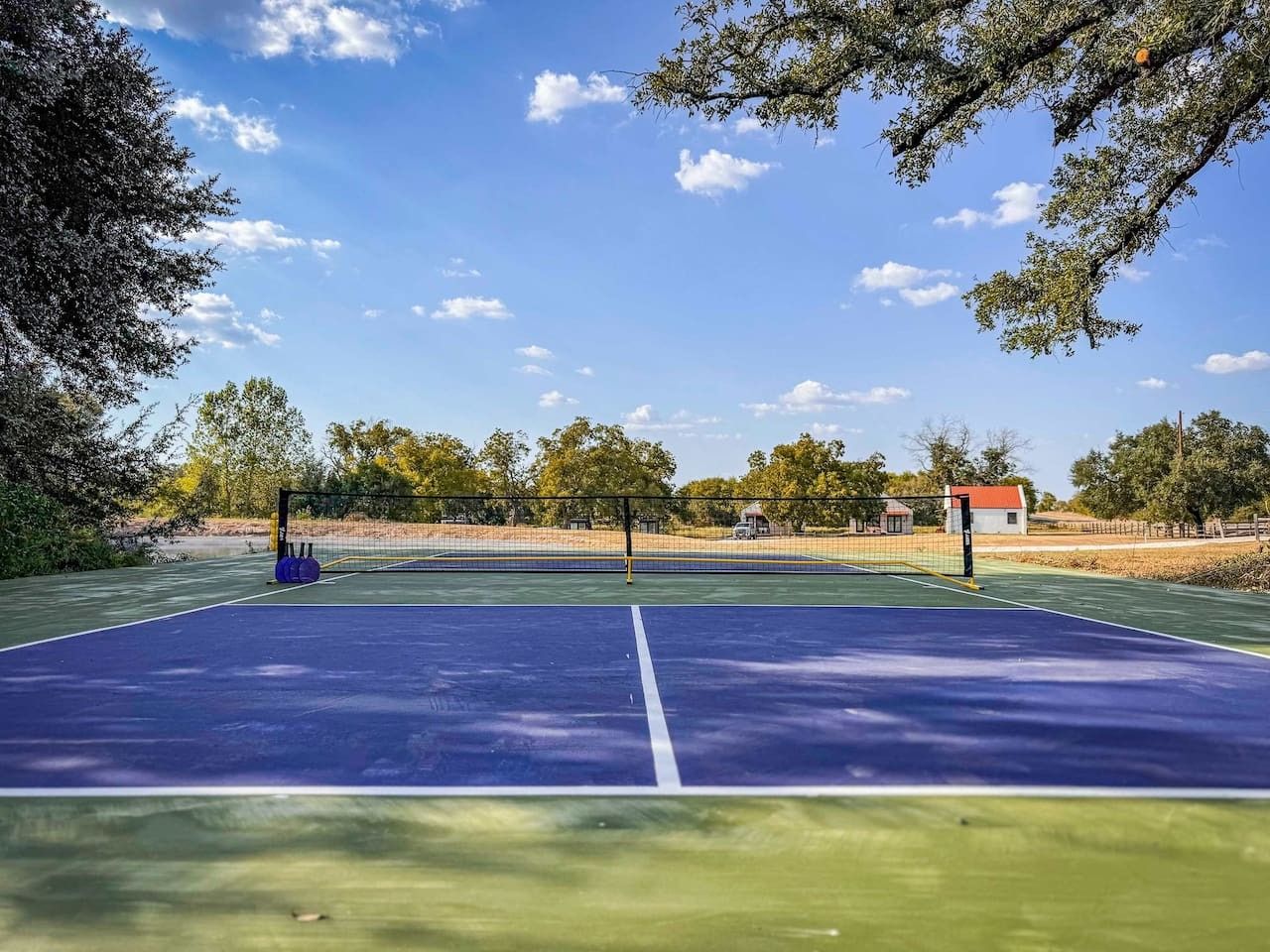 Ein Tennisplatz im Freien mit Netz, umgeben von Bäumen und einem Gebäude im Hintergrund.