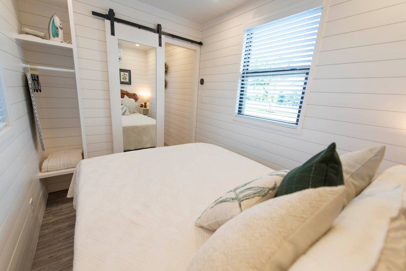 Een slaapkamer met een bed, een raam met jaloezieën, een schuifdeur en een plank met spullen.