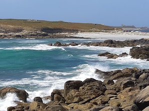 A rocky coastline with waves crashing against the shore, a sandy beach, and a grassy hill in the background. - Beach house within walking distance (Plouguerneau)