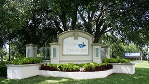 Placa indicando Lake Fairways, com um logotipo com tema aquático, rodeada por vegetação e um cercado branco.