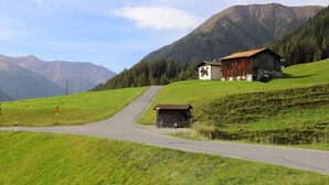 Uma estrada rural leva a um pequeno edifício com telhado vermelho, cercado por campos verdes exuberantes e montanhas ao fundo.