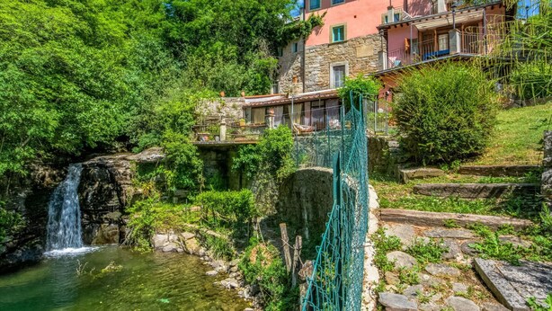 Un edificio de piedra con fachada roja, una cascada pequeña y abundante vegetación.