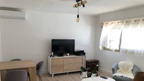 A modern living room with a flat-screen TV, wooden furniture, a white coffee table, and a window with sheer curtains. - 2-room apartment for 2 people close to Moure Rouge beach. (Cannes)
