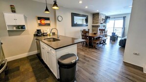 A modern kitchen with a central island, a dining area with a wooden table and chairs, and a view of the living area through a glass door. - Modern Condo, Walk to Gondola, Summer Fun! (Keystone)