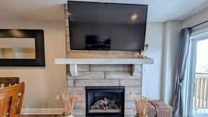 A living room with a fireplace, a television mounted above it, and a dining area with a wooden table and chairs. - Modern Condo, Walk to Gondola, Summer Fun! (Keystone)