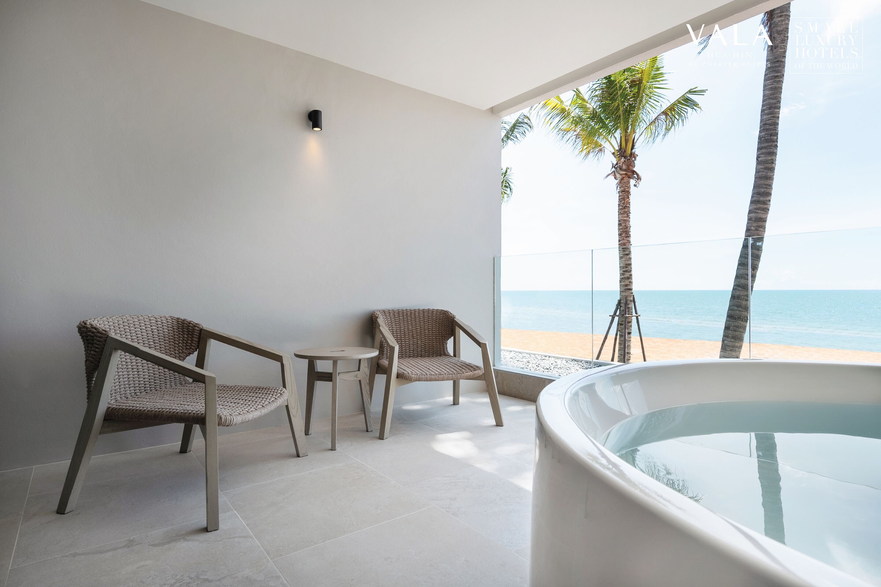 A modern room with two wicker chairs, a small table, and a large bathtub. The room has a view of the beach and palm trees.