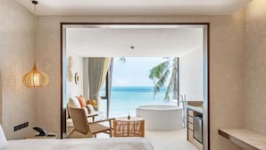 Una habitación de hotel moderna con una cama grande, una zona de estar con vista al océano, una bañera y una mesita de noche con una lámpara.