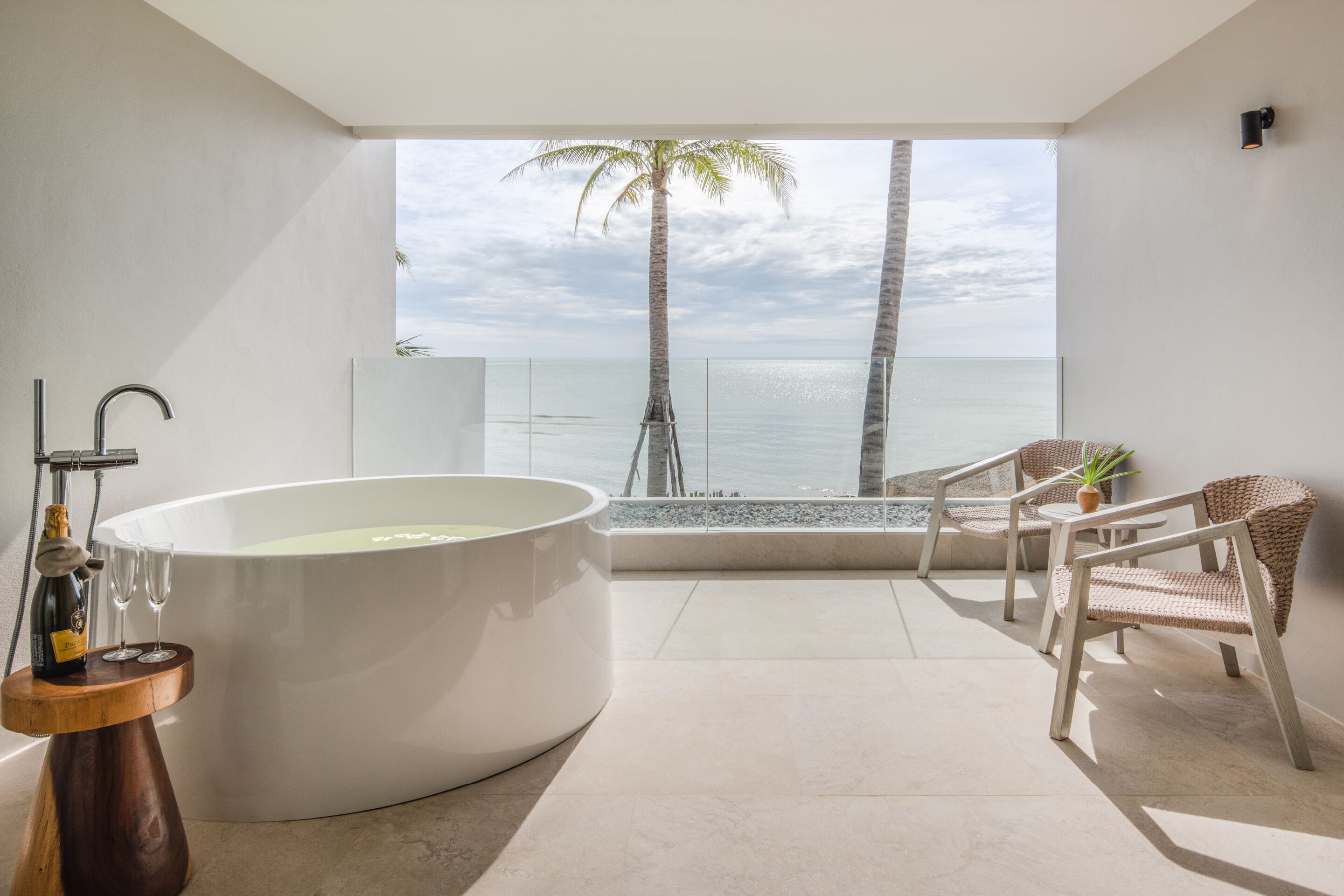 A modern bathroom with a large bathtub, a wooden stool with champagne, and a view of the ocean and palm trees.