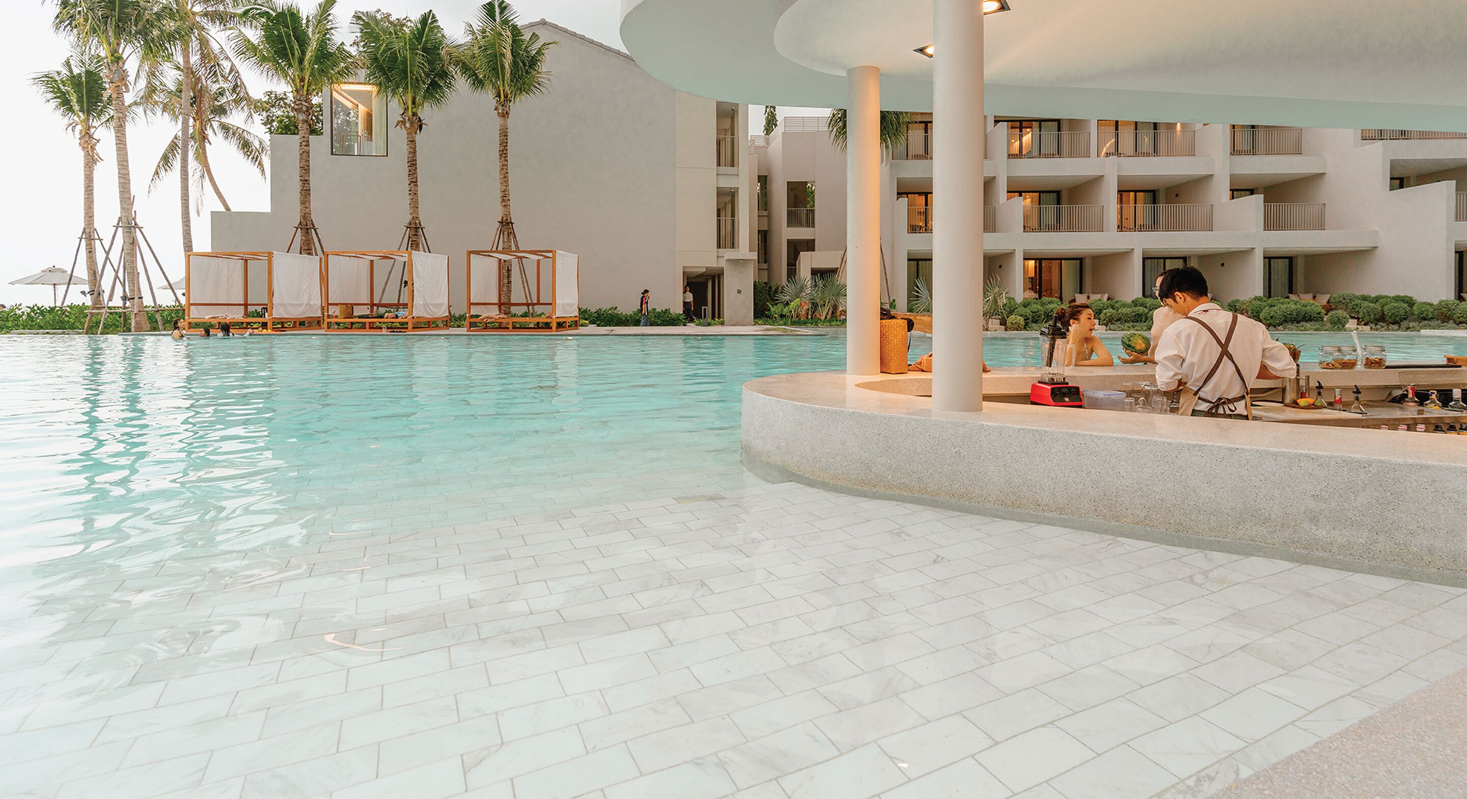 A pool area with lounge chairs, palm trees, and a modern building with balconies.