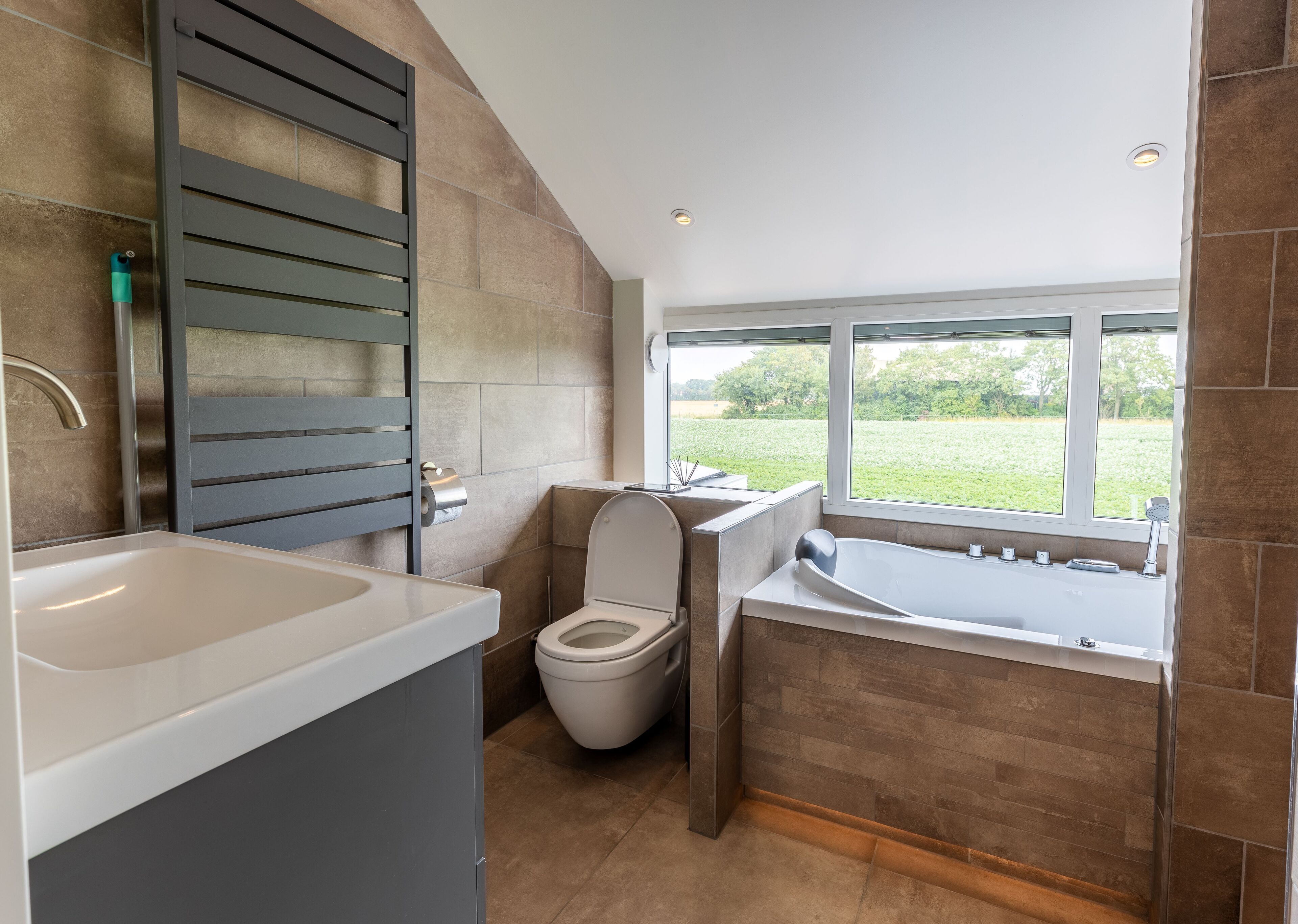 A modern bathroom with a toilet, sink, and bathtub, featuring a large window with a view of greenery.