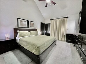 Un dormitorio con cama, ventilador de techo, mesita de noche, cómoda y una puerta que da a otra habitación.