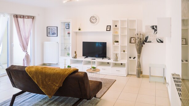 Un salon moderne doté d’un téléviseur à écran plat, d’étagères blanches, d’un canapé recouvert d’un plaid marron et d’une grande fenêtre ornée de rideaux légers.