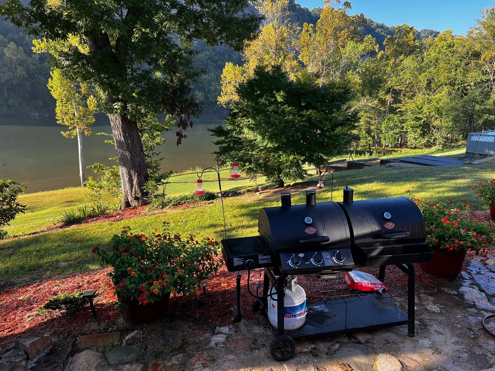 Una parrilla para barbacoa con tanque de propano, rodeada de un jardín con flores rojas y vista a un lago y árboles.