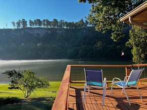 Een houten terras met twee stoelen met uitzicht op een rustig meer, omgeven door bomen en een gazon.