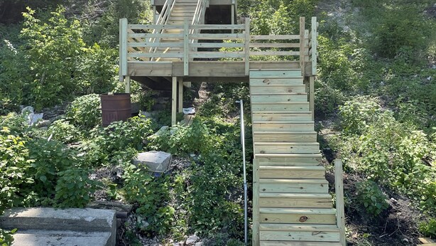Una escalera de madera que sube a una plataforma con rampa, rodeada de follaje y árboles.