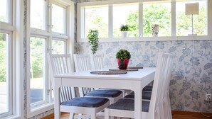 Comedor con una mesa y sillas blancas, una planta en maceta y una ventana con vistas a árboles.