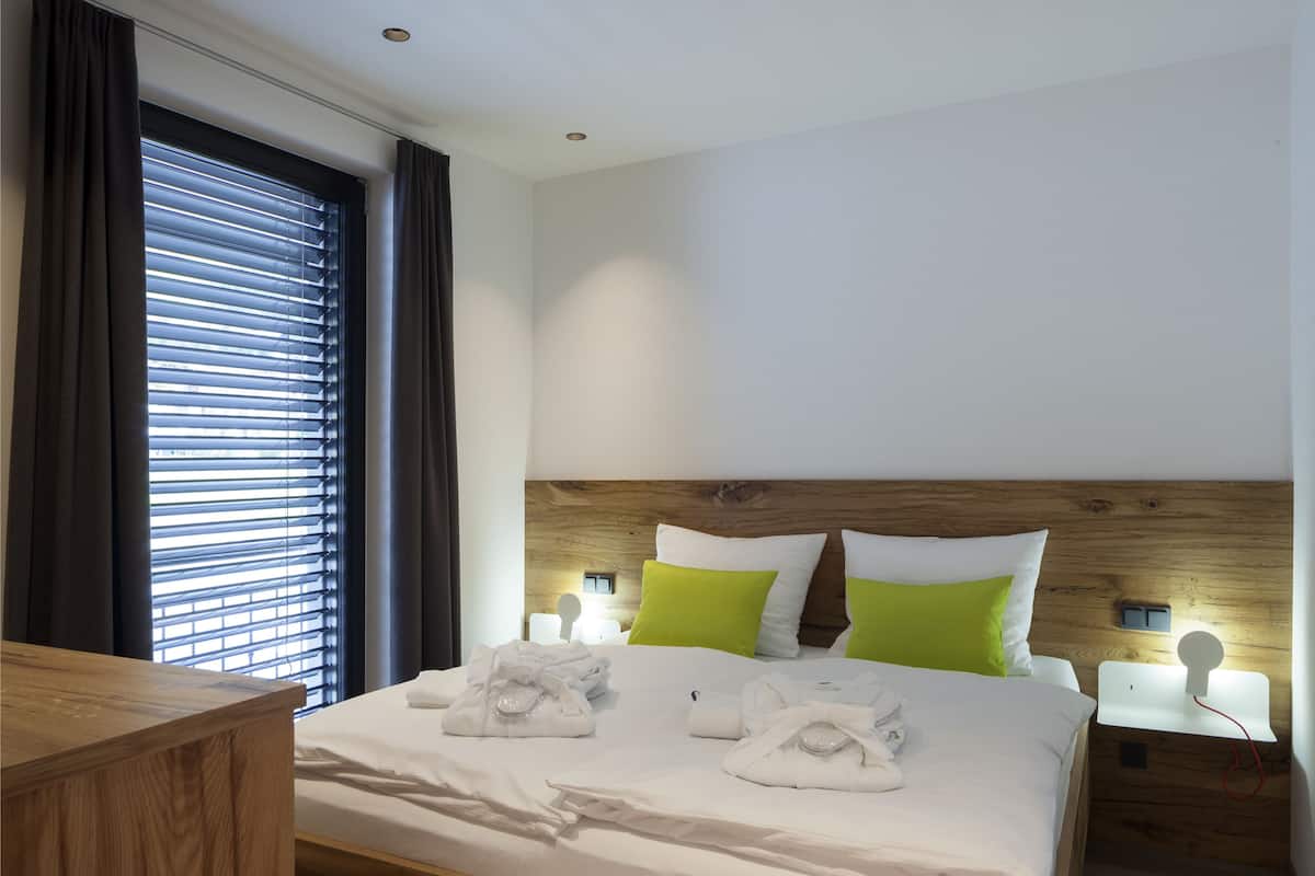 Una cama bien tendida con sábanas blancas y almohadas verdes, un cabecero de madera y una mesita de noche con una lámpara.