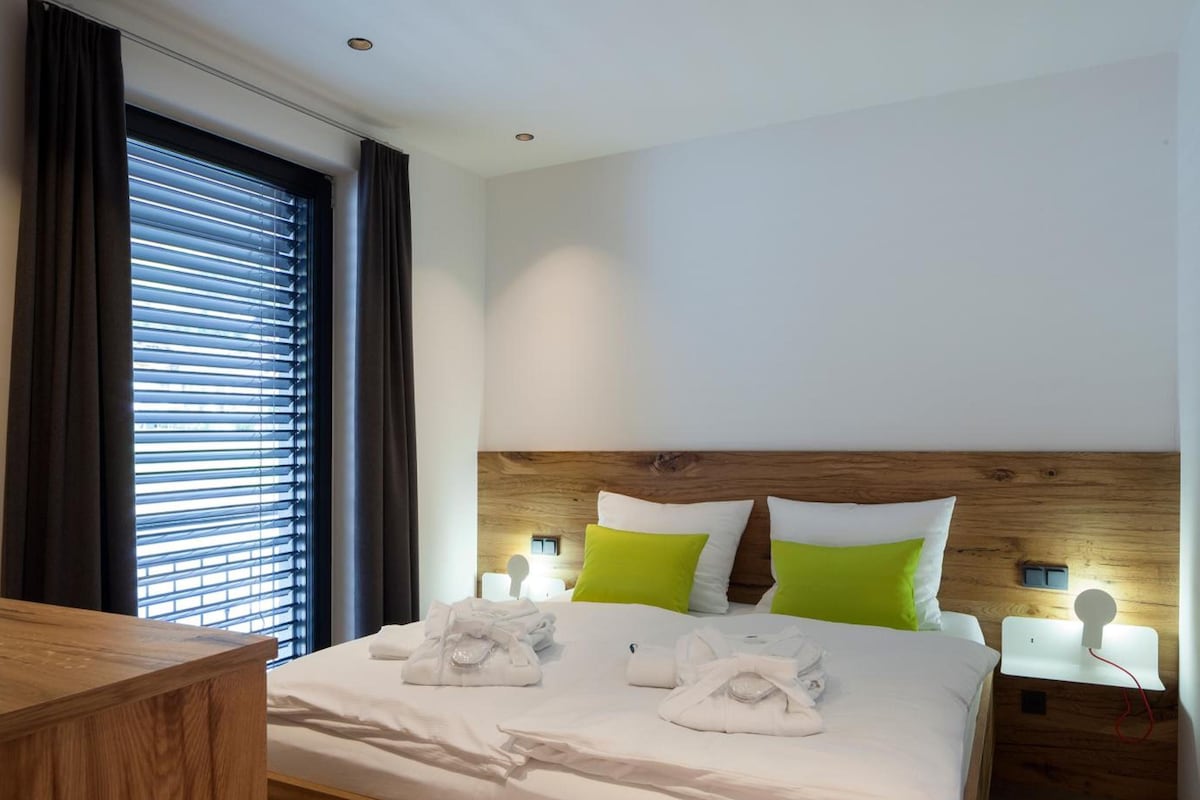 A neatly made bed with white linens and two green pillows, a wooden headboard, and a bedside table with a lamp.