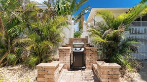 Un espace barbecue extérieur équipé d’un gril à gaz, entouré de palmiers et bordé d’un chemin en pierre.