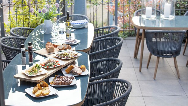 An outdoor dining area with wicker chairs and a glass table set with plates of food, glasses, and candles.