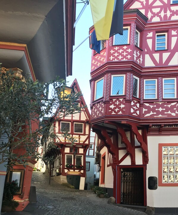 Uma rua estreita de cal çada, flanqueada por casas tradicionais de enxaimel, cada uma com desenhos distintos nas janelas e uma placa indicando um restaurante.
