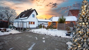 Un quartier résidentiel où le sol est recouvert de neige, une maison blanche avec un toit en pente et un sapin de Noël illuminé.