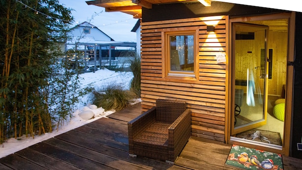 Une terrasse en bois avec une chaise en osier et un banc en bois, une porte en bois avec des panneaux de verre, et un paysage enneigé en arrière-plan.