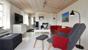 Uma sala de estar moderna com teto de madeira, sofá vermelho, poltrona cinza, um banquinho preto e mesa de centro de vidro.