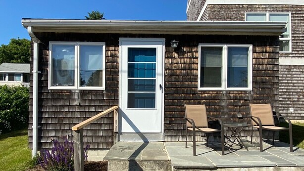 A single-story house with a front door, two windows, a small porch with a table and chairs, and a stone staircase leading to the door.