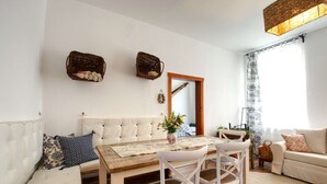 En hyggelig stue med en hvid sofa, et træspisebord og hvide stole. Der er to fletkurve på væggen, et spejl og et vindue med gardiner.