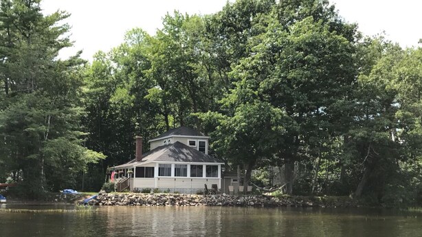A lakeside cabin with a deck, surrounded by dense trees. - Lake Front Taylor Pond Cottage (Auburn)