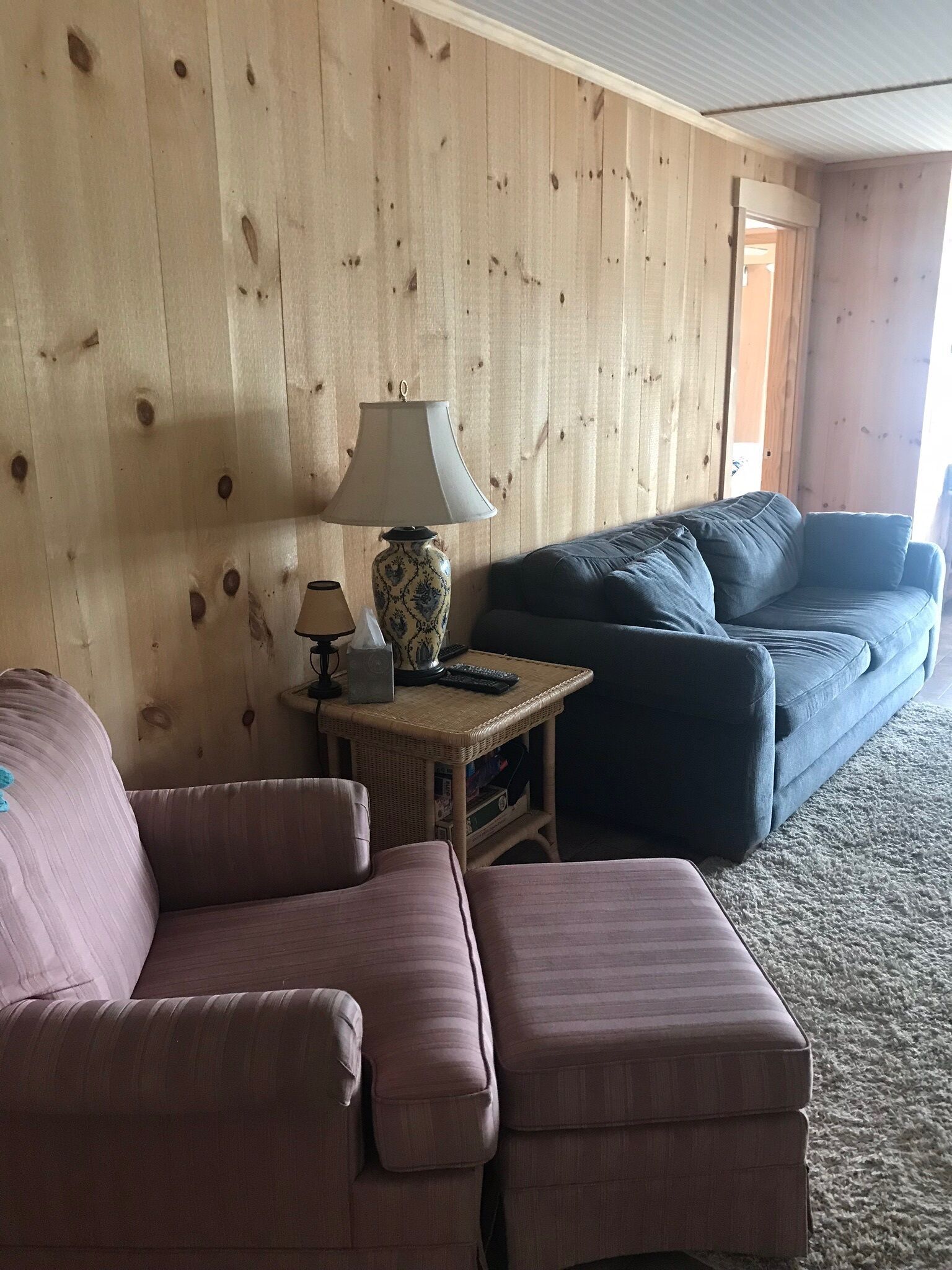 A cozy living room with a wooden wall, a blue sofa, a pink sofa, and a small table with a lamp and remote controls.