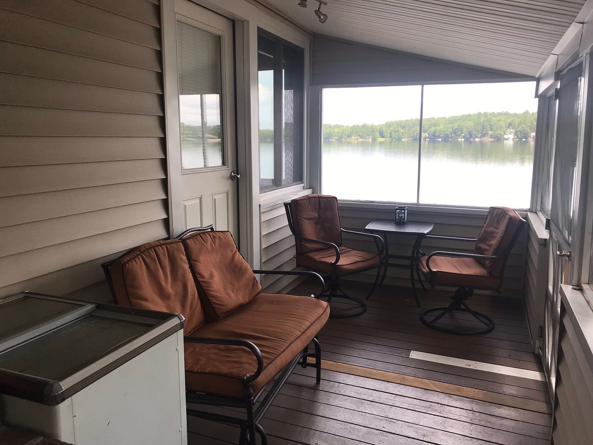 A covered porch with a brown leather sofa and two chairs, a small table, and a view of a lake.