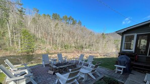 Un patio con sillas Adirondack y una chimenea de piedra, con vista a un río y un área boscosa.