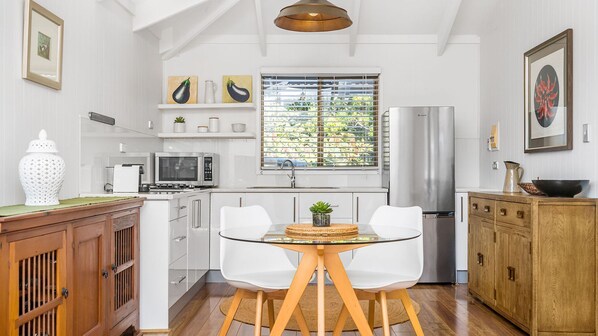 Une cuisine moderne avec des meubles blancs, un réfrigérateur en acier inoxydable, un four à micro-ondes et un évier. On y trouve une fenêtre avec des stores, une petite table avec des chaises et un meuble en bois.