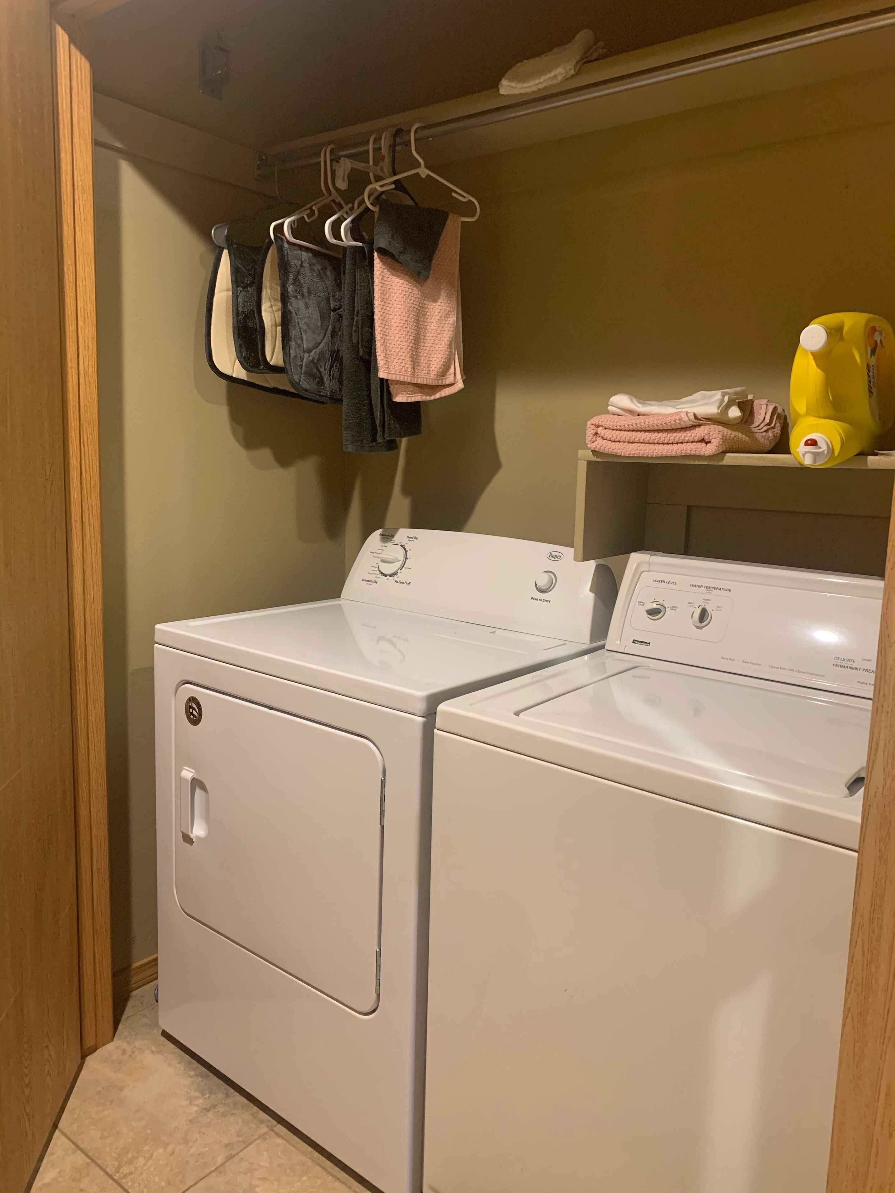 Une buanderie équipée d’un lave-linge et d’un sèche-linge à chargement frontal, d’une étagère avec des serviettes pliées et d’une bouteille jaune de lessive.