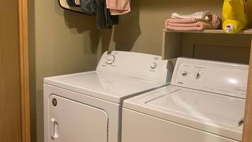 Une buanderie équipée d’un lave-linge et d’un sèche-linge à chargement frontal, d’une étagère avec des serviettes pliées et d’une bouteille jaune de lessive.