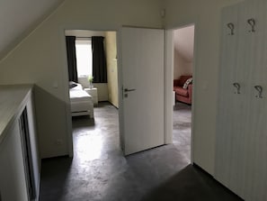 Una habitación con cama, una mesita de noche pequeña, un sofá rojo y una puerta que da a otra estancia.