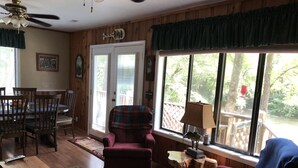 A cozy living room with wooden floors, a dining table with chairs, a plaid armchair, and a view of a deck and trees through large windows. - Country River Cabin (Murphy)