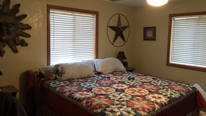 A bedroom with a bed, two windows with blinds, a ceiling fan, a wall decoration, and a small table with a lamp. - Country River Cabin (Murphy)