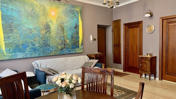 Een eetgedeelte met een ronde tafel, stoelen, een vaas met bloemen, een groot abstract schilderij aan de muur en een kroonluchter.
