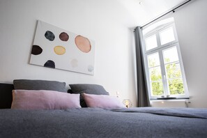 Une chambre à coucher avec un lit, des oreillers, une œuvre d’art murale et une fenêtre avec des rideaux.