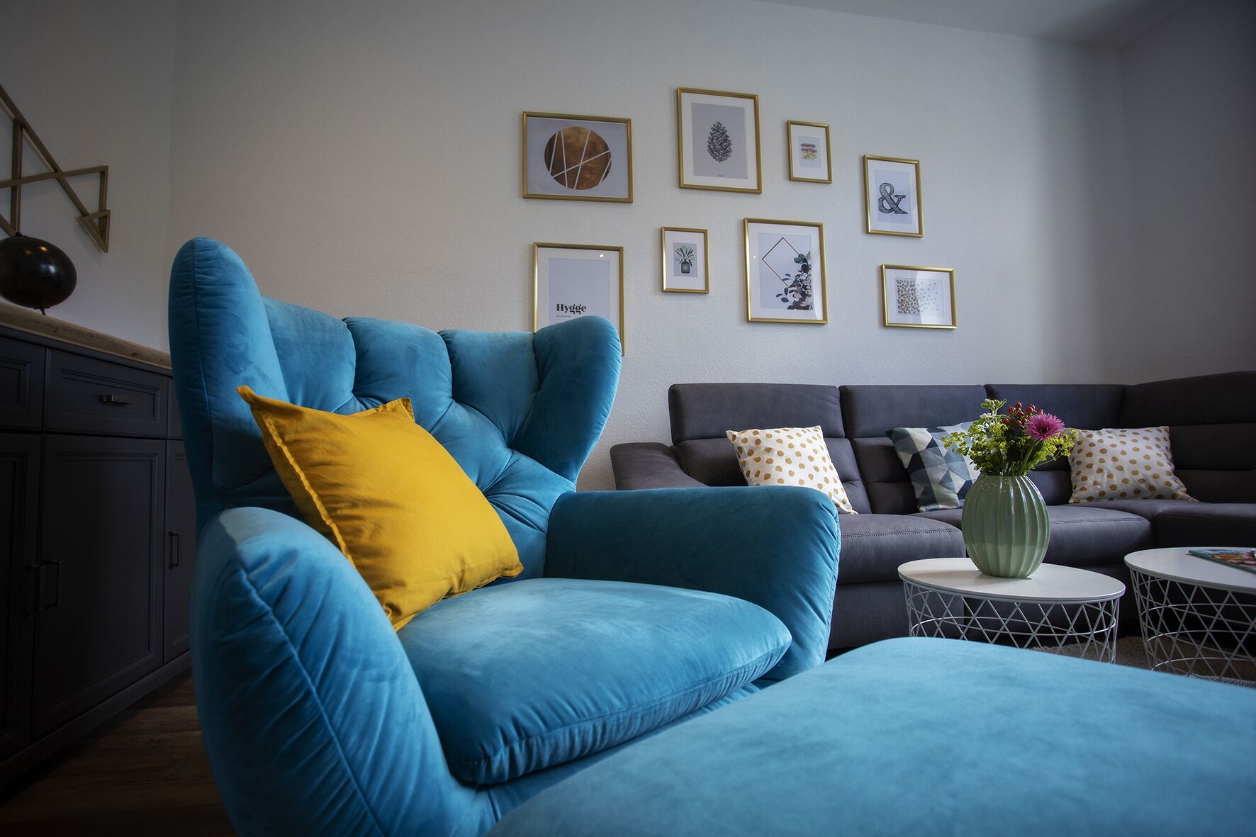 A modern living room with a blue armchair, a grey sofa, and a small round table with a vase of flowers.
