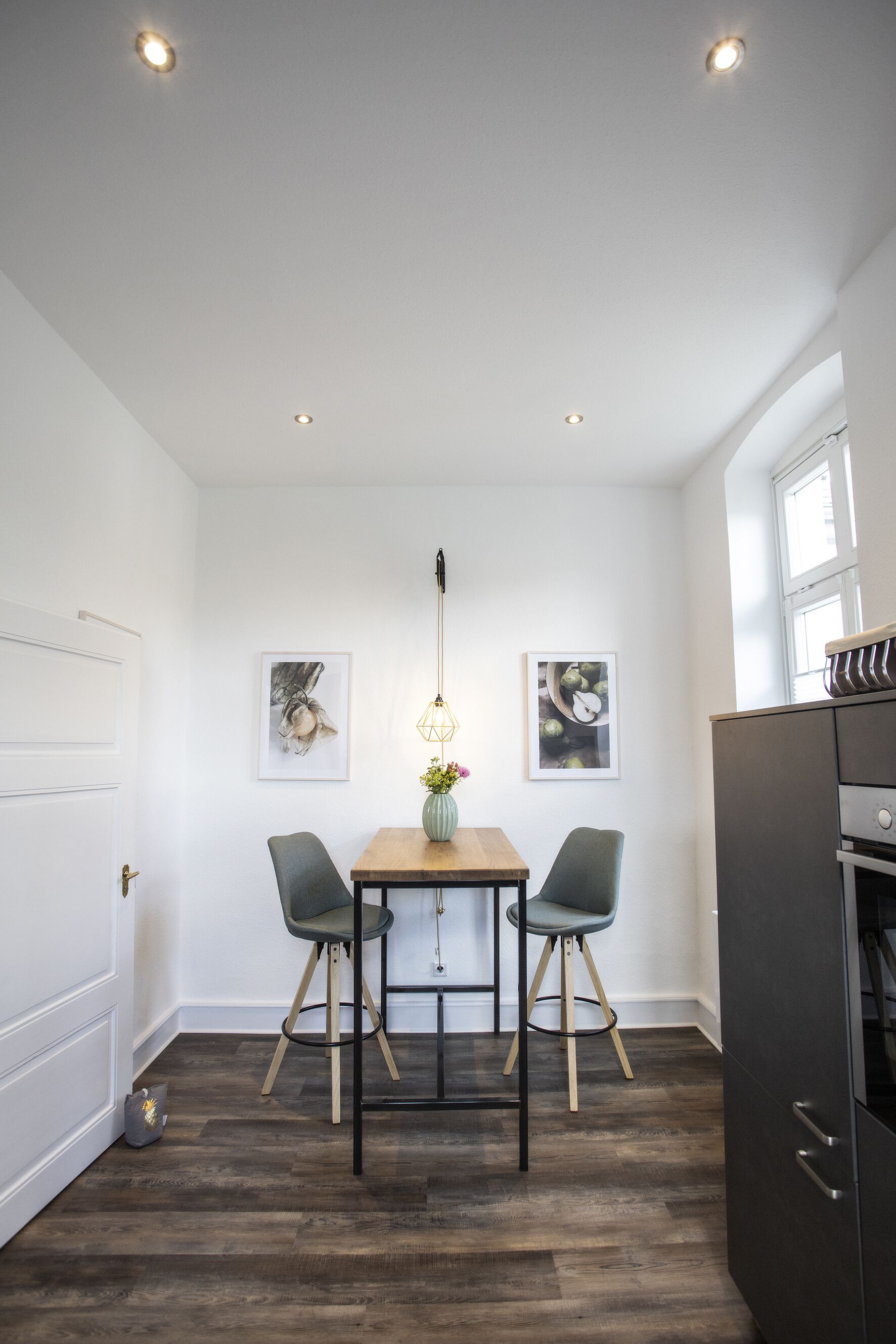 A dining area with a wooden table, two chairs, a vase with flowers, and ceiling lights.