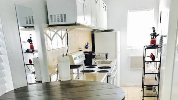 A modern kitchen with white cabinets, a stove, and a sink. There is a round table with two black stools. - Petrina Apartments (Pompano Beach)