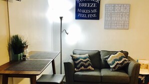 A living room with a dark grey sofa, two patterned cushions, a small table with a plant, and a TV mounted on the wall. - Petrina Apartments (Pompano Beach)