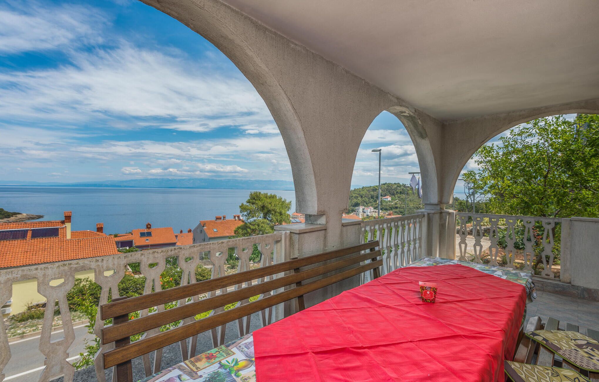 A balcony with a red tablecloth, wooden bench, and a view of the sea and buildings.